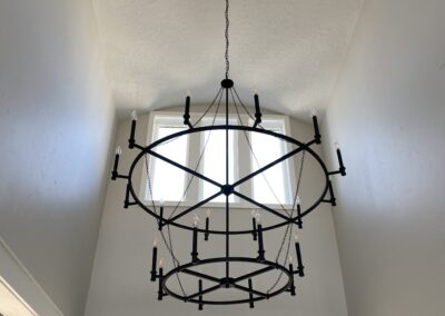 A large, black two-tiered circular chandelier with unlit bulbs hangs from a high ceiling in a bright entryway with tall windows above a dark arched doorway.