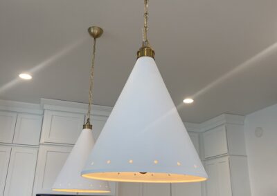Two white cone-shaped pendant lights with brass accents hang from the ceiling in a modern kitchen with white cabinets and a stainless steel refrigerator.