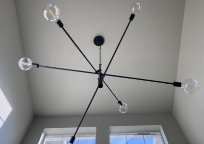 A modern, black sputnik chandelier with five exposed bulbs is mounted on a white ceiling above two large windows letting in daylight.