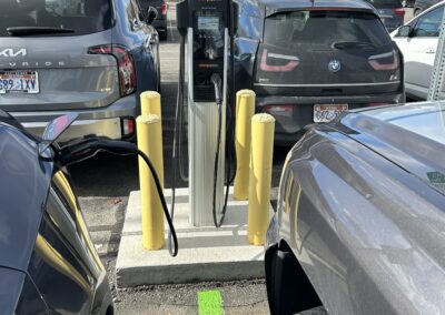 An electric vehicle charging station is situated between parked cars in a parking lot, with yellow bollards surrounding the charger.