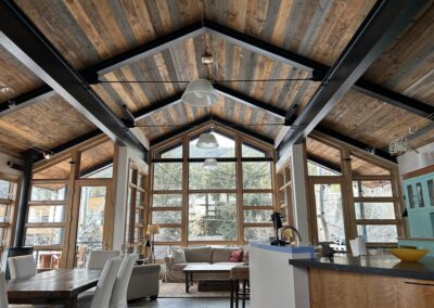 Modern, open-concept living room with vaulted wooden ceiling, large windows, a beige sofa, dining table, and kitchen island overlooking a forested outdoor view.