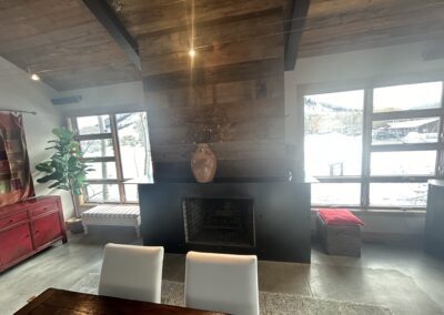A modern living room with a wooden accent wall, central fireplace, large vase, two white chairs, and large windows showing a snowy outdoor scene.