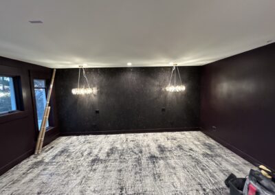 A modern, empty room with dark walls, two hanging chandeliers, a ladder by the window, and a light-colored area rug covering the floor.
