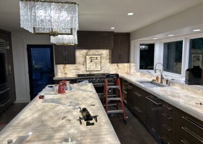 Modern kitchen under renovation with a lit stone countertop, dark cabinets, a step ladder, tools, and appliances visible; overhead crystal light fixture and large windows present.