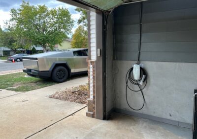 A stainless steel electric pickup truck is parked in a driveway outside an open garage with an electric vehicle charging unit mounted on the wall.